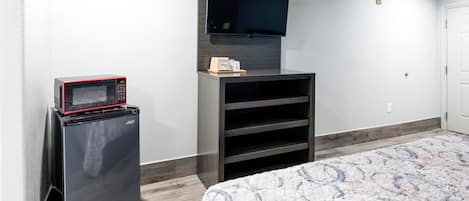 A compact room with a bed, a small refrigerator, a flat-screen TV mounted on the wall, and a built-in shelving unit.