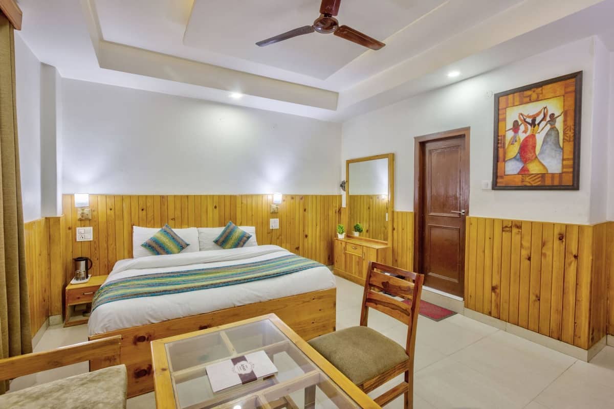 Una habitación de hotel con cabecera tapizada en madera, una cama con ropa de cama a rayas, una mesita de noche de madera, una mesita de noche con superficie de cristal, una silla, un aparador de madera con espejo y un cuadro en la pared.