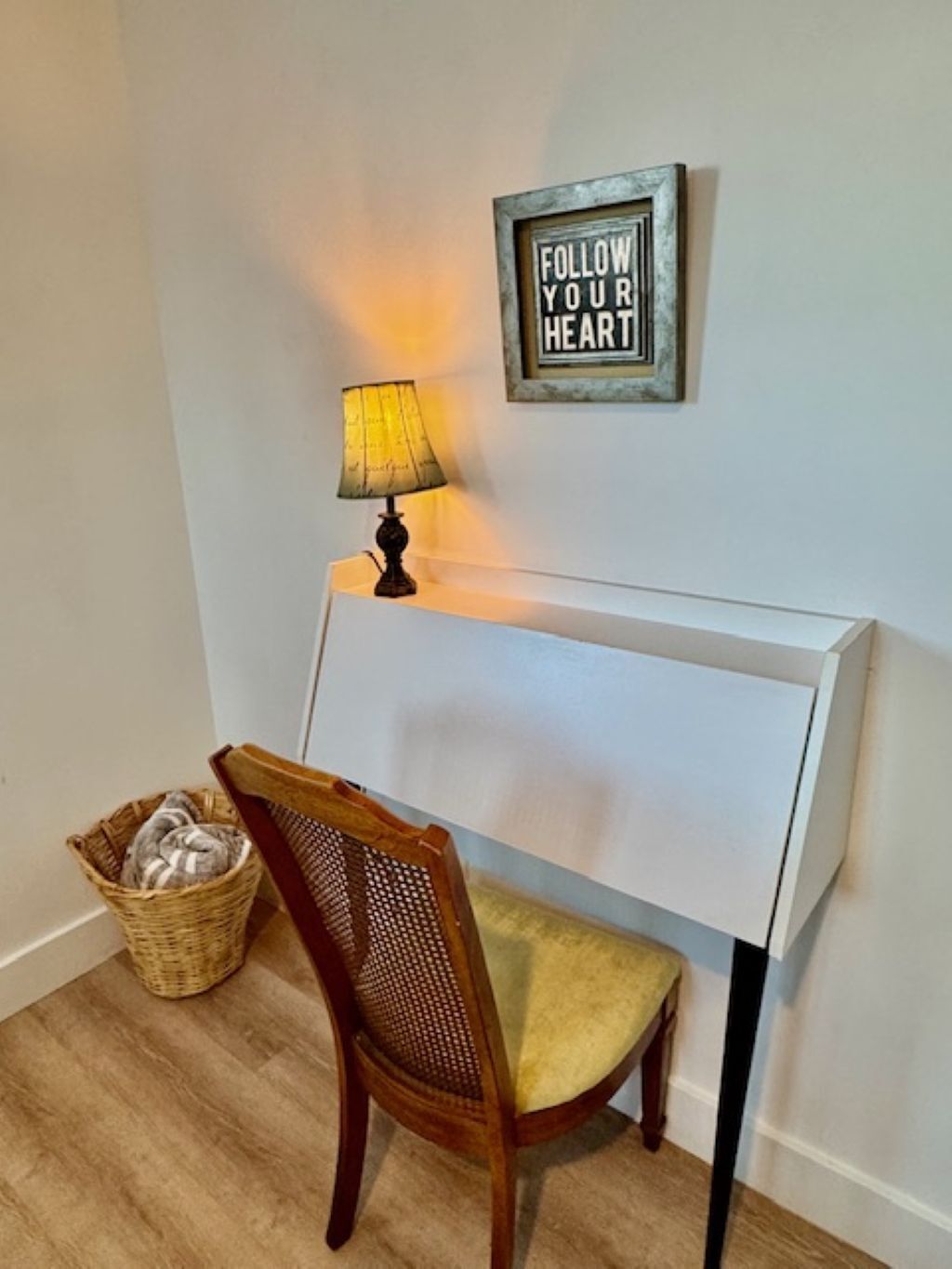 Una habitación con cabecera blanca, una silla de madera con cojín, una lámpara, una cesta de mimbre y un cartel en la pared que dice "FOLLOW YOUR HEART".
