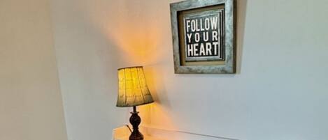 Una habitación con cabecera blanca, una silla de madera con cojín, una lámpara, una cesta de mimbre y un cartel en la pared que dice "FOLLOW YOUR HEART".