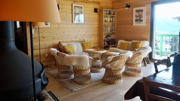 Un interior de madera acogedor, con chimenea, sillones de mimbre, una estantería y vistas a través de un ventanal.