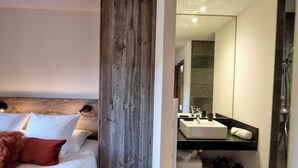 A modern hotel room with a wooden headboard, a large bed with pillows, a bedside table, and a bathroom with a sink, mirror, and shower. - Loft Odette (Font-Romeu-Odeillo-Via)