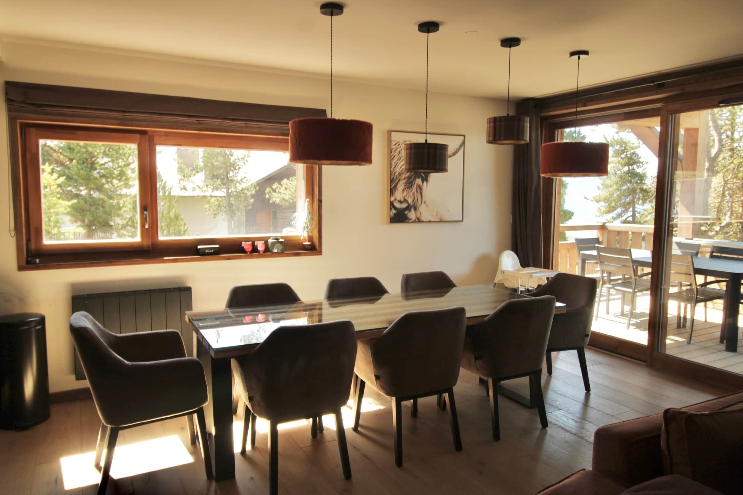 A dining area with a glass table, chairs, pendant lights, and a view through large windows.
