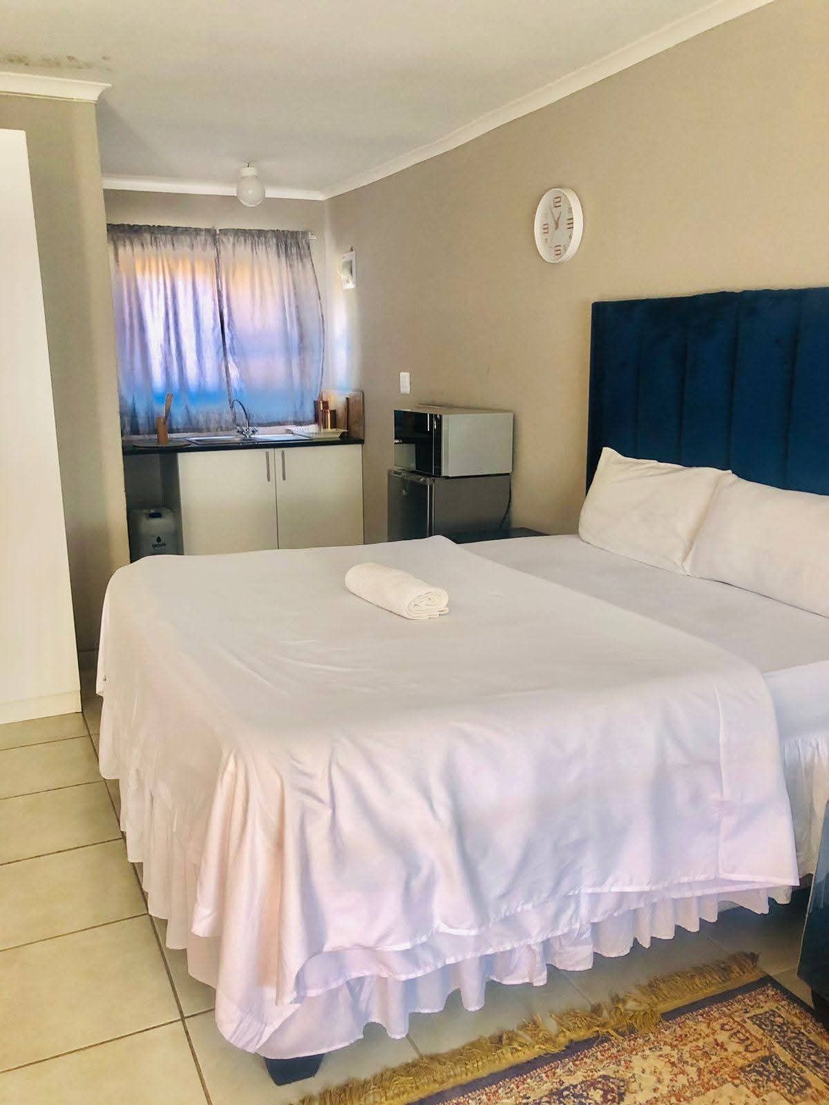A hotel room with a large bed, a small kitchenette, a window with curtains, and a clock on the wall.