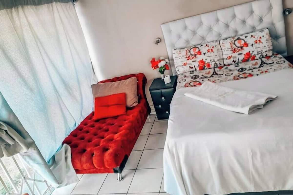 Een slaapkamer met een groot bed, een rode, knopachtige bank, een nachtkastje en een raam met gordijnen.