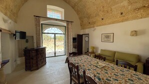 Une pièce comprenant un lit, une commode, un téléviseur, une table à manger avec des chaises, et une vue sur l’extérieur par une fenêtre avec des rideaux.