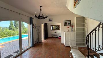 Une pièce spacieuse dotée d’une grande porte-fenêtre coulissante donnant sur une piscine, d’un lustre, d’une console blanche et d’un escalier avec une rampe en métal.