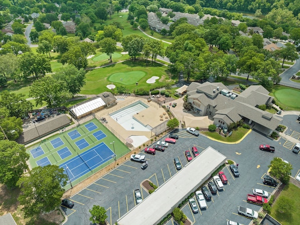 Vista aérea de um campo de golfe com quadra de tênis e piscina, cercado por casas residenciais e um estacionamento.