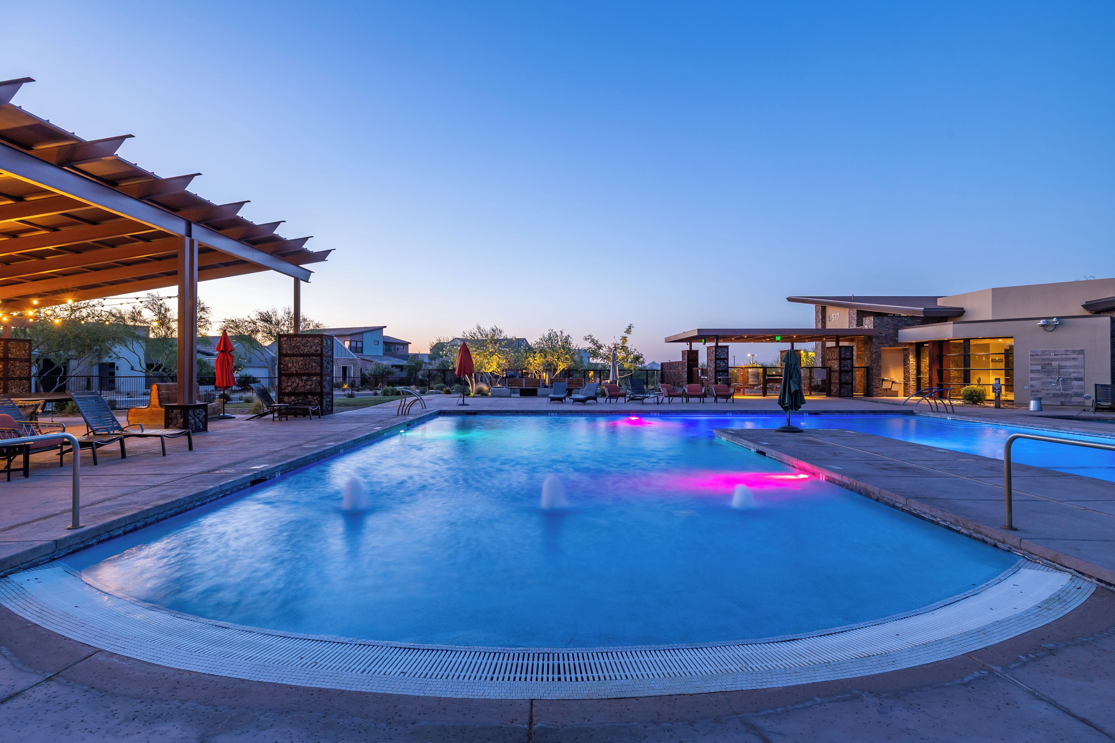 A swimming pool with a pergola and lounge chairs, surrounded by a landscaped area with a building in the background.