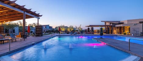 Una piscina con una pérgola y sillas de descanso, rodeada de jardines y un edificio al fondo.
