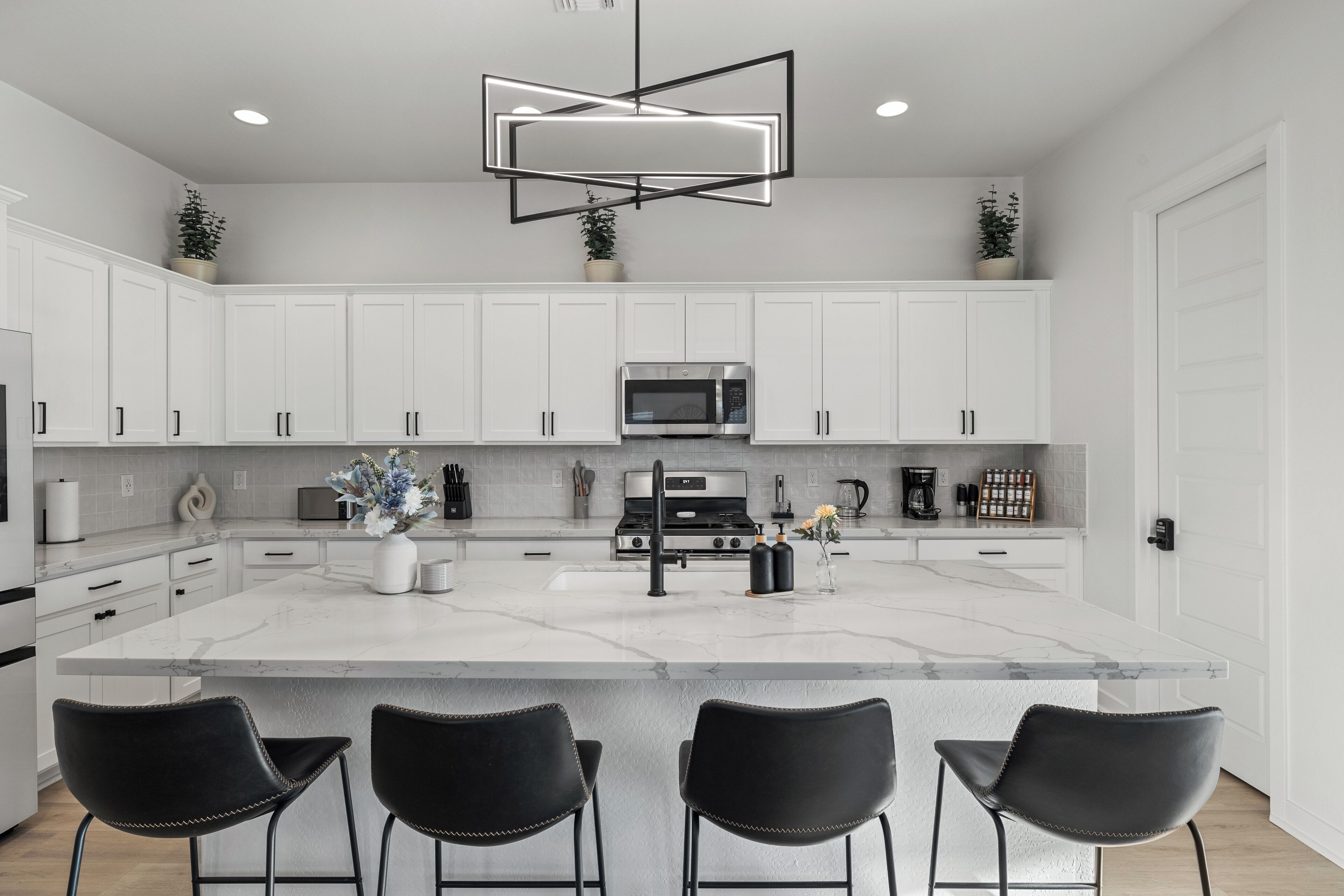 A modern kitchen with white cabinetry, a central island with bar stools, a stainless steel microwave, and a black pendant light.