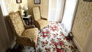 Una habitación con dos sillones con estampado, una mesita con lámpara, alfombra floral y papel tapiz decorado, además de una ventana con contraventanas.