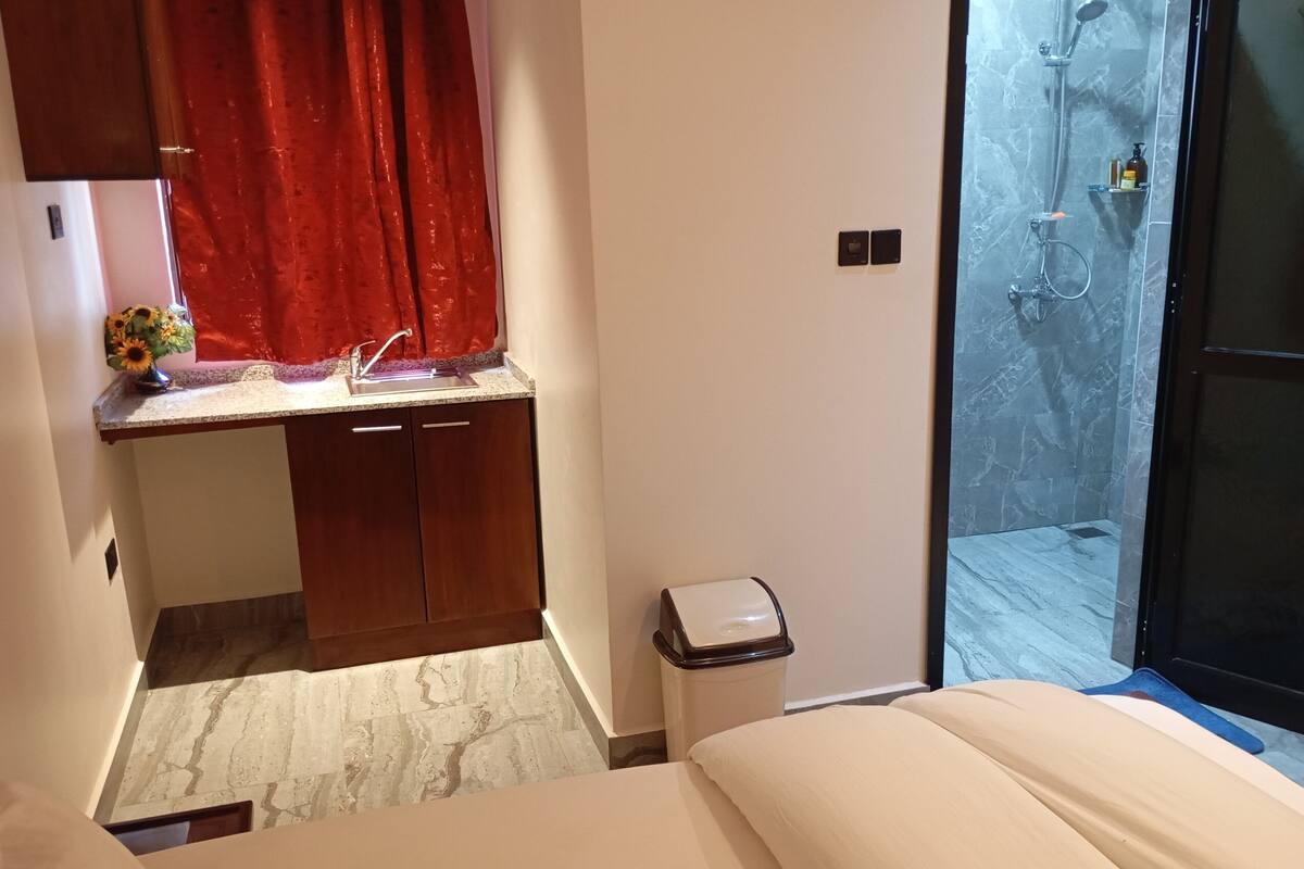 A bedroom with a bed, a small desk with a sink, a trash bin, and a shower area.