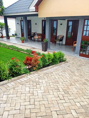 Uma área de estar coberta ao ar livre, com cadeiras e mesas, um caminho pavimentado e um jardim com diversas plantas e flores.