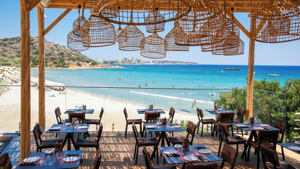 An outdoor dining area with tables and chairs overlooking a beach and the sea. - The River Suites - Adults Only (Agios Nikolaos)