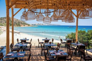An outdoor dining area with tables and chairs overlooking a beach and the sea. - The River Suites - Adults Only (Agios Nikolaos)