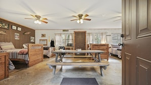 A rustic cabin interior with wooden furniture, a central picnic table, ceiling fans, and a door leading outside.