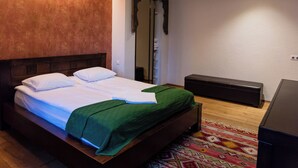 Una cama bien tendida con sábanas blancas y un edredón verde, un cabecero de madera, un banco oscuro y un tapete estampado en el suelo.