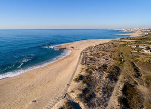 Un paysage côtier avec une plage de sable, une mer bleue et limpide, et une silhouette de ville qui se dessine à l’horizon.