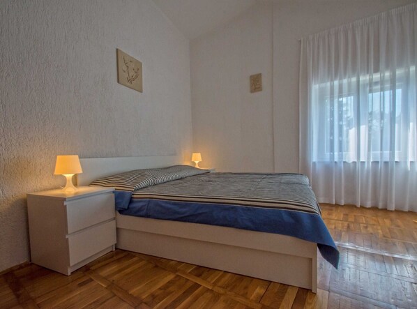 Un dormitorio con cama, dos lámparas de noche, una mesita de noche y una ventana con cortinas traslúcidas.