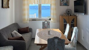 A living room with a sofa, a dining table with chairs, a TV, and a view of the sea through a window.