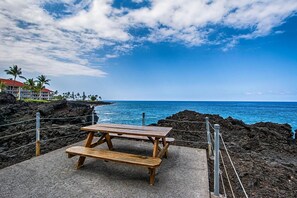 Ein Holztisch für ein Picknick auf einer Betonplatte mit Blick auf das Meer und ein Gebäude mit rotem Dach im Hintergrund.