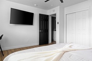 A bedroom with a bed, a TV mounted on the wall, and a door leading to a bathroom. - Private Pool + Arcade • 8BR Group Retreat • Sleeps 18 (Mesa)