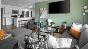 A modern living room with a grey sofa, a glass coffee table, and a flat-screen TV mounted on a green wall.
