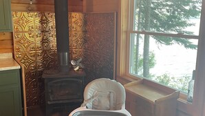 A cozy cabin interior with a wooden interior, a child’s high chair, a wood-burning stove, and a window overlooking a forest. - Cozy cottage in charming Lincoln, ME with AC (Lincoln)