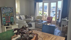 A spacious living room with a large window, a dining table with a basket of plants, and a view of the marina. - 1st line water front 5 Minutes to the beach and 10 mins to old town center. (Hellevoetsluis)