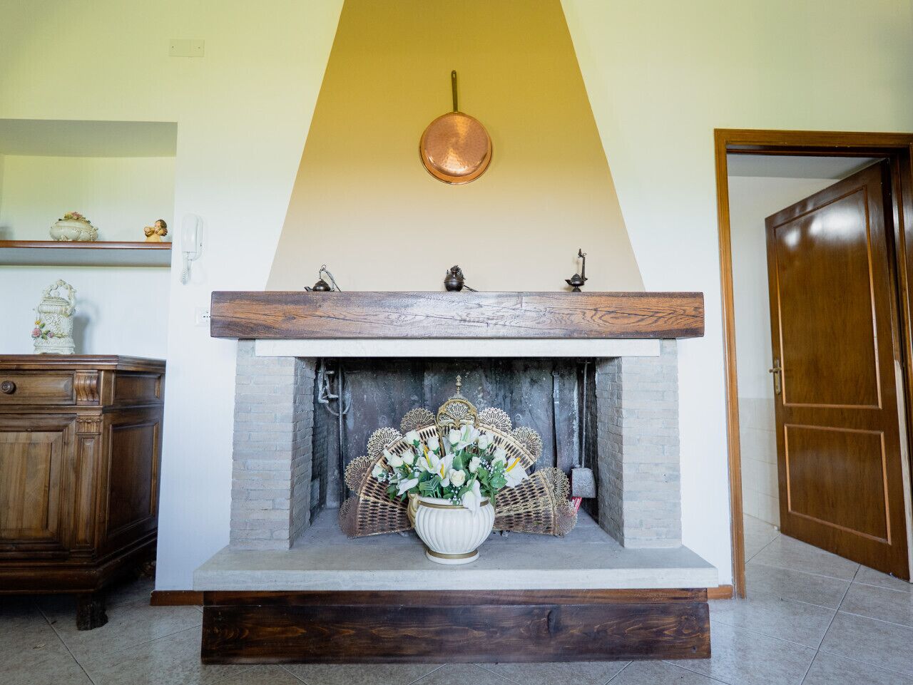 A fireplace with a white vase of flowers, a wooden mantel, and a copper pot hanging above.