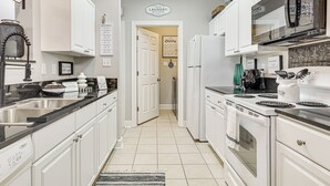 Una cocina con gabinetes blancos, un lavamanos de acero inoxidable, una estufa y un refrigerador. Hay carteles que indican la cocina y el cuarto de lavado.