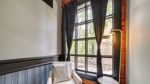 Una habitación con un sillón blanco, una mesita redonda pequeña, una ventana con cortinas negras y una lámpara fijada a la pared.