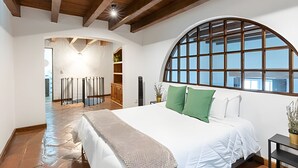 A bedroom with a large bed, a wooden ceiling, a large arched window, and a view of a cityscape.