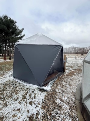 A geodesic dome tent with a metal frame and a dark fabric exterior, set on a gravel surface with sparse snow.