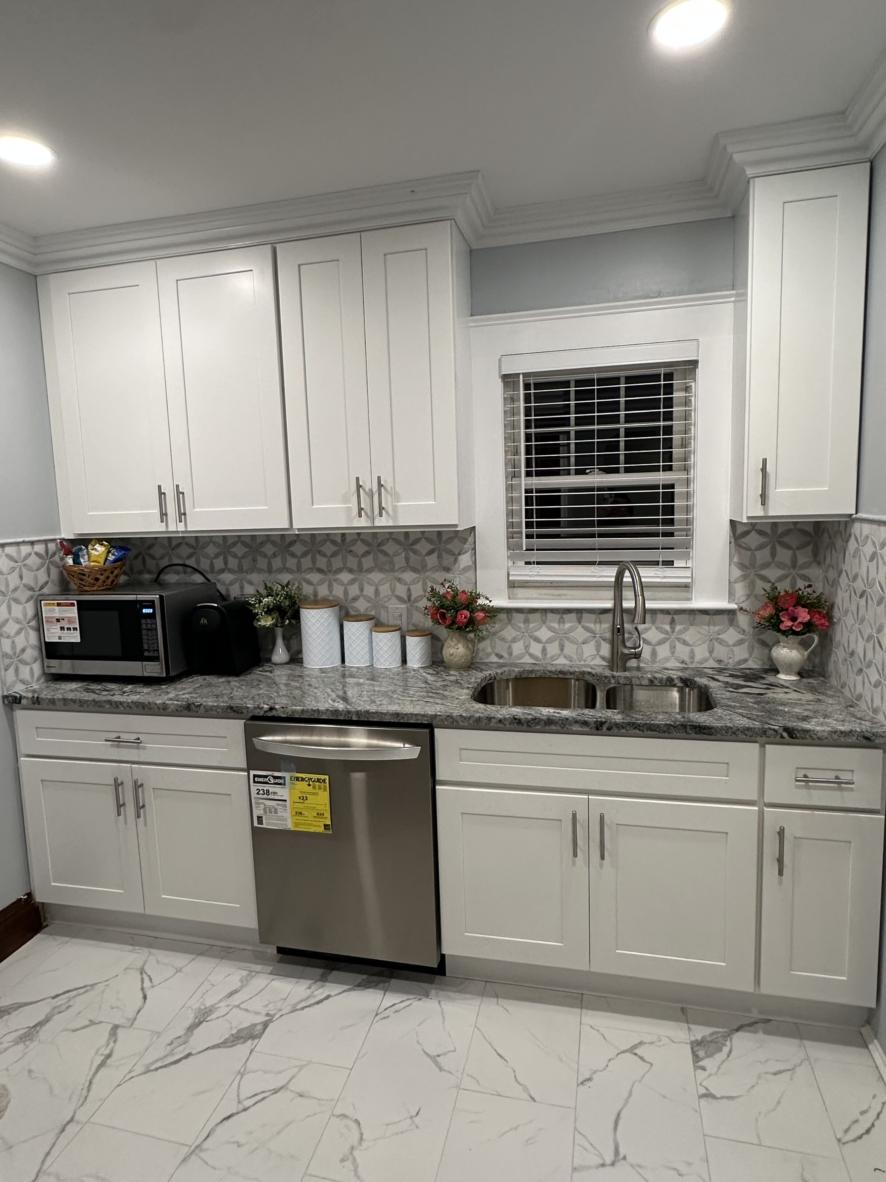 A kitchen with white cabinets, a stainless steel dishwasher, a microwave, and a window with blinds.