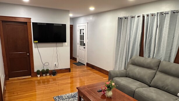 Uma sala de estar com piso de madeira, um sofá cinza, uma mesa de centro com um vaso e uma TV de tela plana na parede.