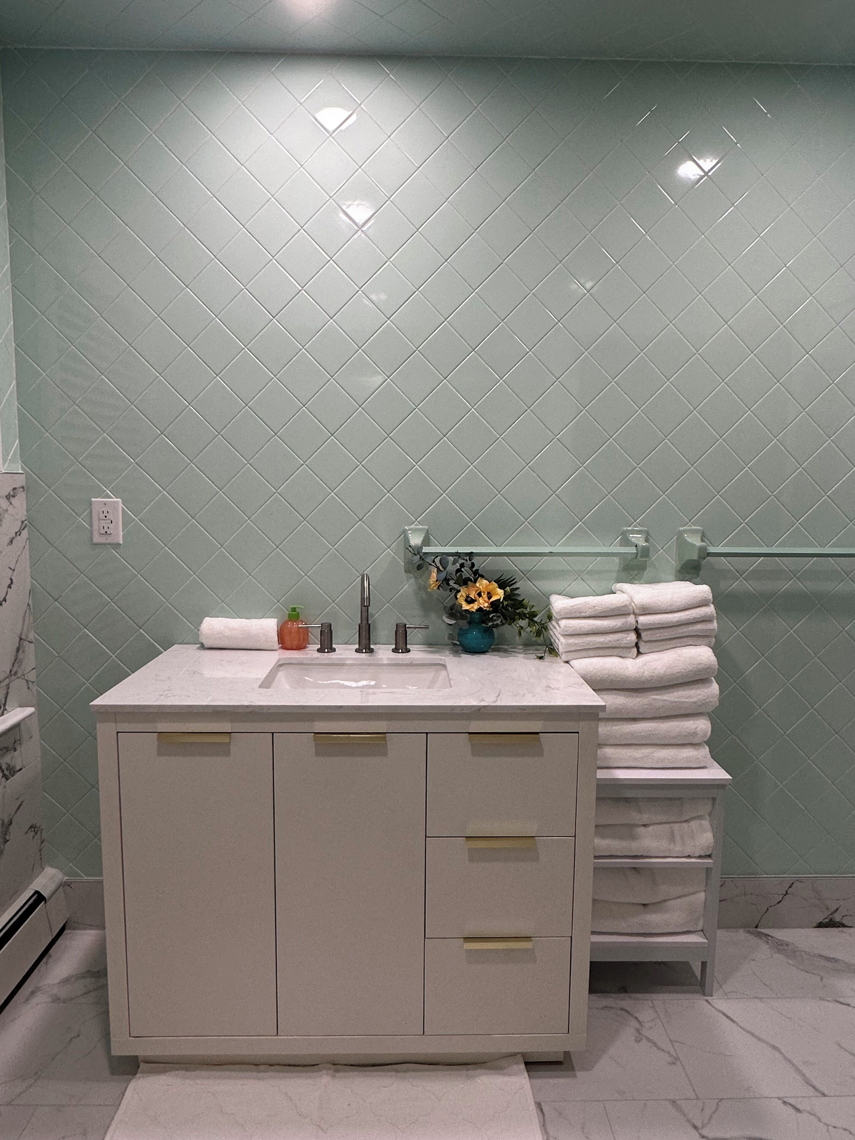 Un baño con tocador blanco, lavamanos y espejo. Hay un perchero con toallas dobladas y un florero con flores en la encimera. La pared está revestida con un patrón de diamantes.