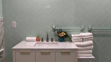 Un baño con tocador blanco, lavamanos y espejo. Hay un perchero con toallas dobladas y un florero con flores en la encimera. La pared está revestida con un patrón de diamantes.