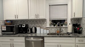 Uma cozinha moderna com armários brancos, lava-louças de aço inoxidável, micro-ondas, cafeteira e pia com janela acima.