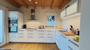 Una cocina moderna con armarios blancos, una isla central, hornos empotrados, un ventanal y techo de madera.