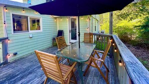 A wooden deck with a glass table and chairs, surrounded by a light green house with windows and string lights.