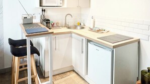 A compact kitchen with a microwave, sink, and a small refrigerator. There is a wooden stool and a table with a laptop and some items on it.