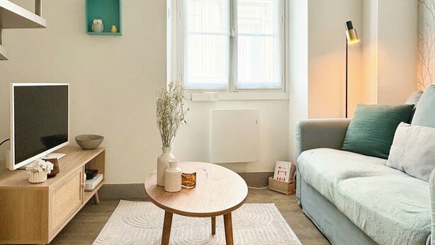 A modern living room with a round wooden table, a sofa, a TV, and a window with blinds.