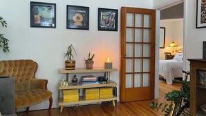 Una sala de estar con un sillón capitoné, una consola de madera con plantas y libros, una puerta con paneles de vidrio esmerilado y una cama al fondo.