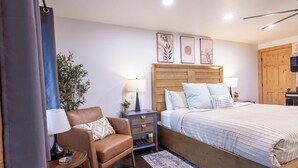 A bedroom with a wooden headboard, a bed with striped bedding, a nightstand with a lamp, a leather chair, and a small plant.