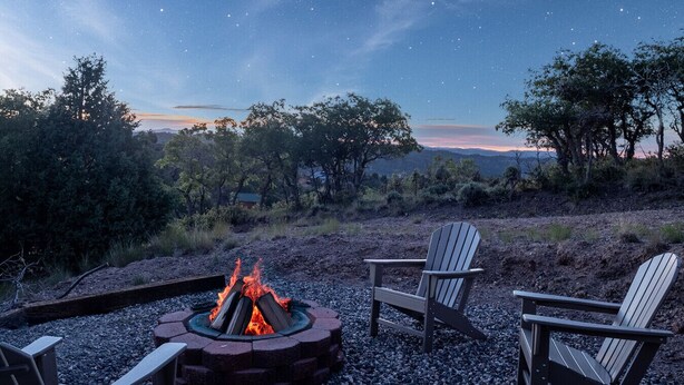 A fire pit with two Adirondack chairs in a natural setting with trees and a starry sky.