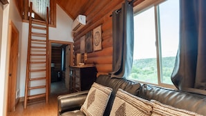 Ein Ferienhäuschen-Interieur mit Holzbalkendecke, einer Ledersofa und einer Leiter, sowie einem Fenster mit Vorhängen.