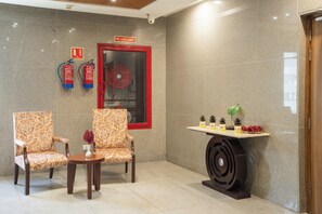 A lobby with two patterned chairs, a small table, a decorative table with pinecones, and a fire hose cabinet. - AR Suites KLJ Square (Gurugram)