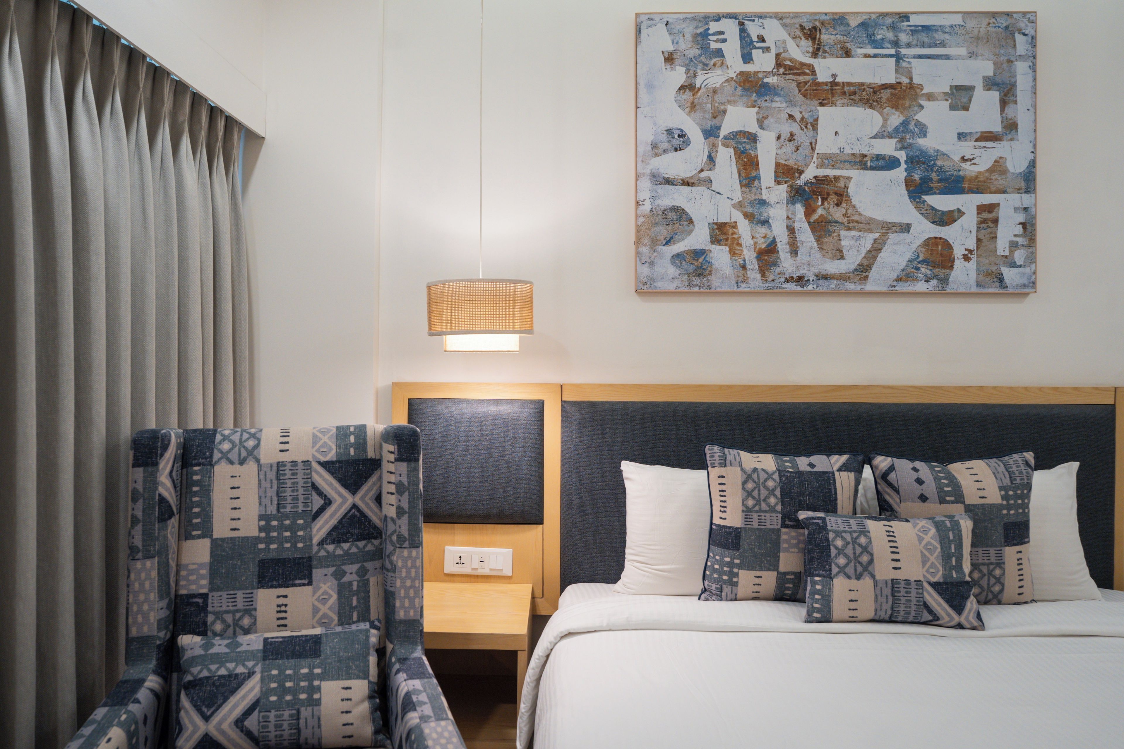 A hotel room with a bed, a nightstand, a lamp, a chair, and a wall art piece.